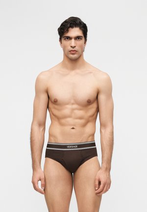 Mees toonuses kehatüübiga, kes kannab pruune Versace aluspükse, seisab ilma mustrita valge tausta ees, näoga otse vaataja poole ja käed lõdvestunud.