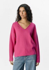 Suéter rosa de cuello en V hecho de un tejido de punto suave, con mangas largas y relajadas y un ajuste ligeramente holgado, combinado con jeans de mezclilla azul.