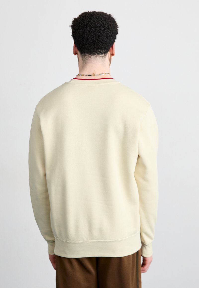 Cremefarbener Sweatshirt mit rundem Halsausschnitt und geripptem Saum, der eine subtile Textur und einen kontrastierenden roten Akzent am Kragen aufweist.