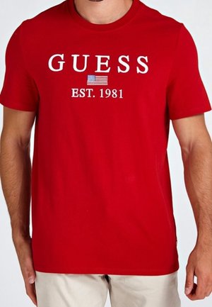 Rode korte mouwen t-shirt met "GUESS," een Amerikaanse vlag en "EST. 1981" op de voorkant gedrukt, gedragen met lichtgekleurde broek.