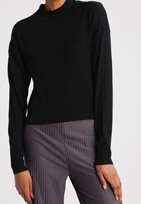 Maglione lungo nero a maniche lunghe con colletto alto, realizzato in morbido tessuto a maglia. Presenta una vestibilità comoda e polsini a costine, abbinato a pantaloni grigi a righe.