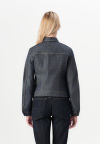Blonde woman facing away wearing a fitted dark denim jacket and matching dark denim jeans against a white background.