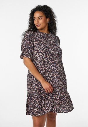 Vrouw met krullend haar die een donkere korte mouwen jurk met bloemenprint draagt, kijkt opzij, staand tegen een effen witte achtergrond.