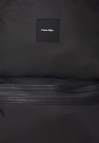 Fekete textúrájú hátizsák sima zipzárral, fekete bőr Calvin Klein téglalap alakú logóval. Minimalista kialakítás.