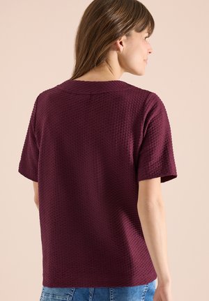Femme portant un haut bordeaux texturé à manches courtes et un jean bleu, tournée de dos sur un fond beige neutre.