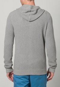 Šedá pletená mikina s texturovaným povrchem, žebrovanými manžetami a lemem. Má šňůrku na kapuci. Nošena přes modré kalhoty.