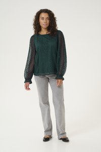 Blusa in pizzo verde scuro con maniche lunghe e voluminose, caratterizzata da un motivo geometrico. Indossata con jeans dritti grigio chiaro e ballerine nere.