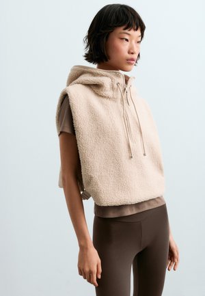 Jeune femme debout de profil, portant un sweat à capuche sans manches en polaire beige sur un t-shirt marron à manches courtes et des leggings marron foncé.