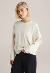 Hanorac bej deschis cu decolteu rotund și mâneci raglan. Textură moale, croială lejeră, asortat cu pantaloni negri cu talie înaltă.