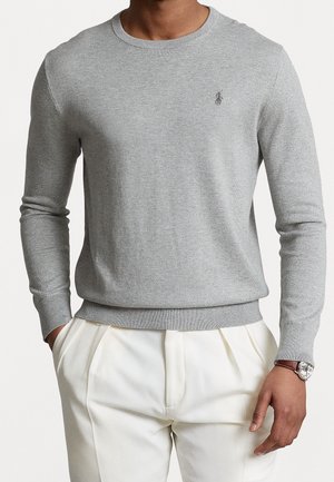 Mann trägt einen hellgrauen Strickpullover mit langen Ärmeln und weiße plissierte Hose, Uhr am linken Handgelenk, Hände teilweise in den Taschen.