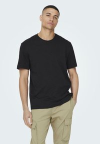 Black cotton t-shirt with short sleeves and a round neck, displayed on a male model. Paired with khaki cargo pants. Simple design, no patterns.