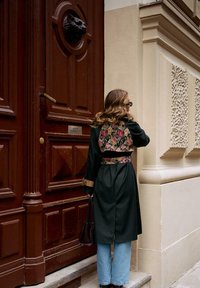 Czarny płaszcz z kwiatową tkaniną z tyłu, złote akcenty na rękawach, w połączeniu z jasnoniebieskimi rozszerzanymi dżinsami i czarną torebką.