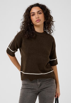 Femme aux cheveux bouclés portant un pull marron à manches courtes avec des rayures blanches et un jean gris, debout avec une expression neutre.