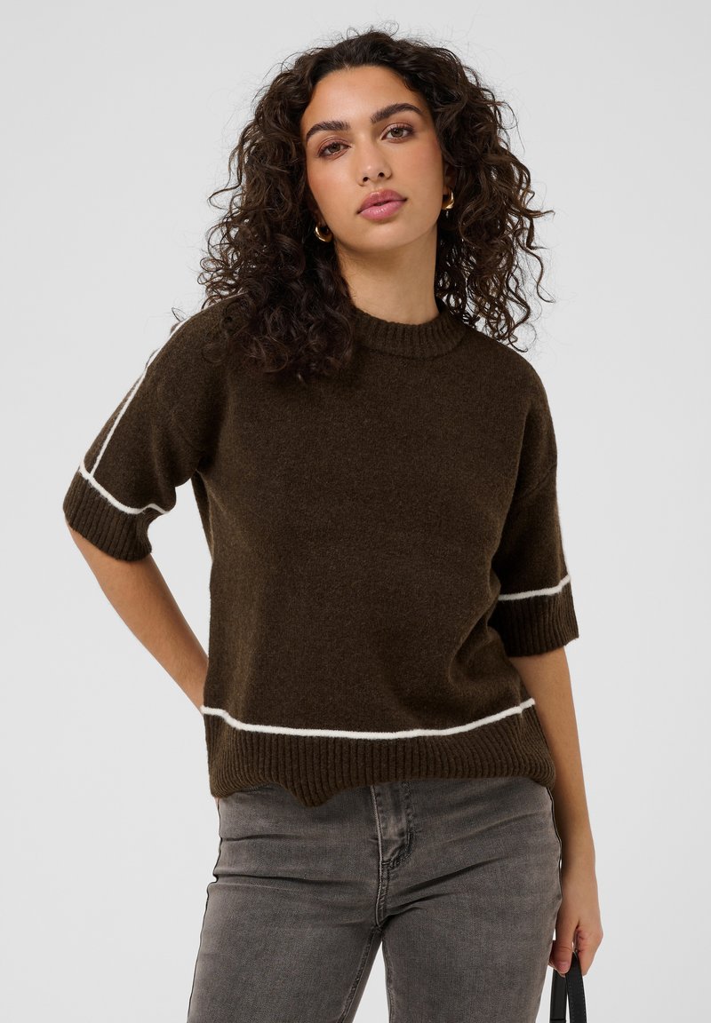 Femme aux cheveux bouclés portant un pull marron à manches courtes avec des rayures blanches et un jean gris, debout avec une expression neutre.