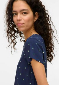 Marineblauwe shirt met korte mouwen, voorzien van een verspreid groen en wit loofpatroon. Gladde stof en ronde halslijn. Krullend haar omlijst het gezicht.