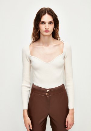 Haut blanc à manches longues côtelé avec un décolleté en V profond, associé à un pantalon marron taille haute avec deux boutons dorés à la taille.