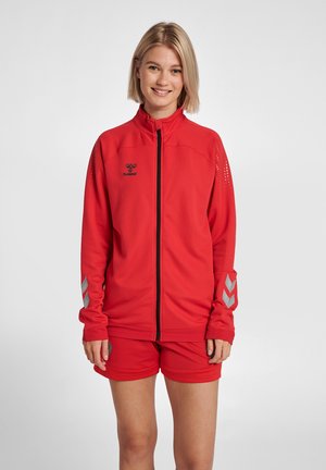 Rote sportliche Zip-Jacke aus strukturiertem Stoff, mit schwarzem Logo und silbernen Chevron-Akzenten an den Ärmeln, kombiniert mit passenden Shorts.
