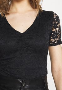 Černý krajkový top s výstřihem do V a krátkými rukávy. Materiál je zdoben květinovými vzory a texturovanými detaily. Nosený s černým koženým páskem.