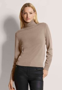 Beige Rollkragenpullover aus weichem, strukturiertem Stoff mit langen Ärmeln und taillierter Silhouette, kombiniert mit schwarzen Slim-Fit-Hosen.