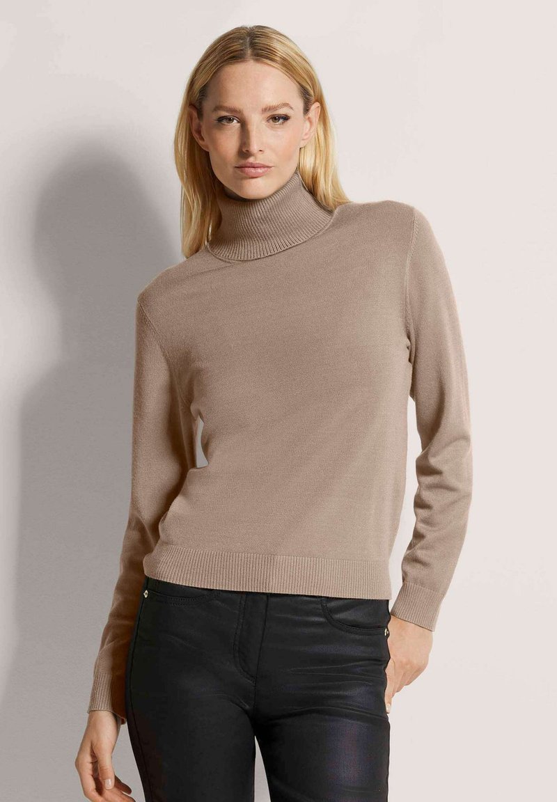 Beige Rollkragenpullover aus weichem, strukturiertem Stoff mit langen Ärmeln und taillierter Silhouette, kombiniert mit schwarzen Slim-Fit-Hosen.