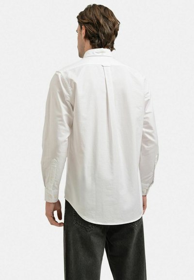 Vit långärmad skjorta med knappmanschetter, ryggsömn och rundad nederkant. Tillverkad av mjukt tyg, bärs med mörka jeans.