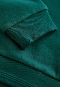 Zelená mikina s textúrovanou tkaninou, rebrovanými manžetami a lemom. Farba je bohatá, tmavozelená, s hladkým povrchom.