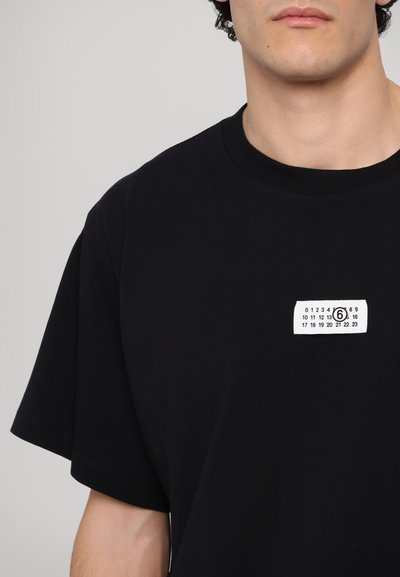 T-shirt noir à manches courtes en coton. Présente une étiquette blanche rectangulaire avec des chiffres et un cercle au centre sur la poitrine.