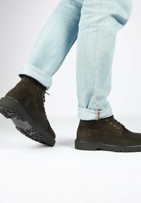 Stivaletti alla caviglia in suede marrone con suole in gomma testurizzata, design con lacci e punta rinforzata. Indossati con jeans azzurri a polsino.