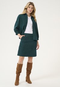 Chemise en velours côtelé teal avec des boutons, assortie à une jupe teal. Bottes en daim marron jusqu'aux genoux. T-shirt blanc en dessous. Texture lisse.