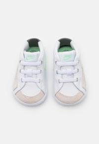 White sneaker with beige suede accents, contrasting black heel, green logo, and textured tongue. Features white laces and rubber sole.