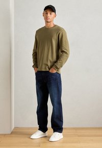 Cămașă lungă cu mâneci în verde măsliniu, cu textură canelată, purtată cu blugi negri din denim și adidași albi. O șapcă neagră adaugă o notă casual.