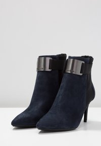 Bottes cheville en daim bleu marine avec un bout pointu, dotées d'une bande métallique et d'un panneau élastique noir pour la flexibilité. Talon stiletto haut.