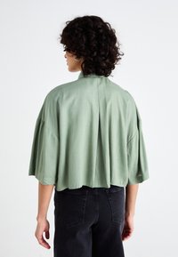 Camisa de mangas curtas, cropped, em tecido verde claro com textura mate e detalhe de pregas nas costas, combinada com calças pretas de cintura alta.