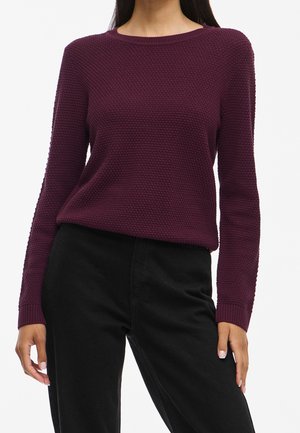 Femme portant un pull prune texturé à manches longues associé à un pantalon noir taille haute sur un fond blanc uni.