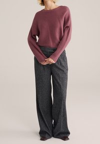 Růžově zbarvený svetr s žebrovanou strukturou a dlouhými rukávy, kombinovaný s tmavě šedými širokými kalhotami s vysokým pasem a vzorem, a tmavě hnědými botami.