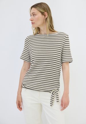 Strikket top med korte ærmer i cremefarve med sorte zigzag-striber. Har en rund halsudskæring og en sidelåse detalje ved kanten.