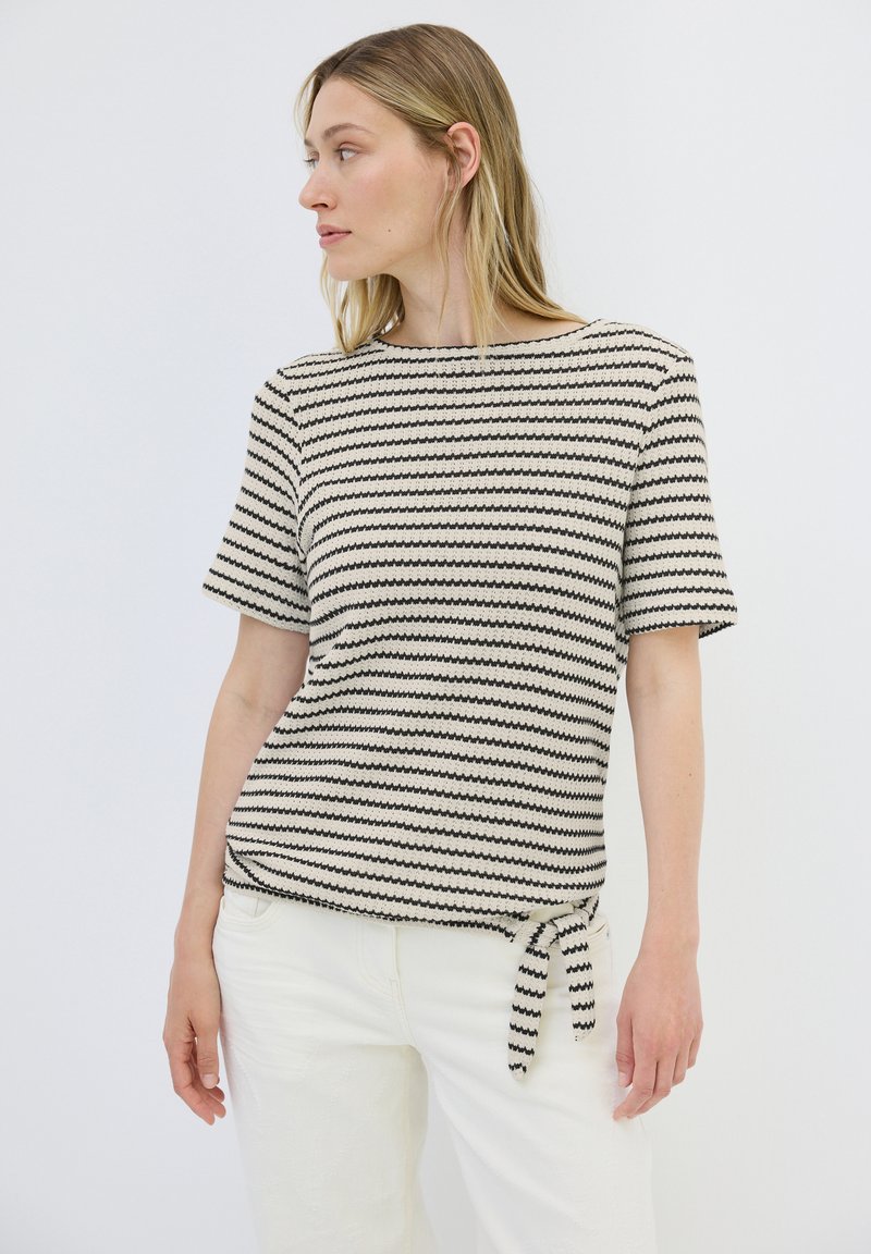 Strickoberteil mit kurzen Ärmeln in Creme mit schwarzen Zickzackstreifen. Mit Rundhalsausschnitt und einem seitlichen Bindeband am Saum.