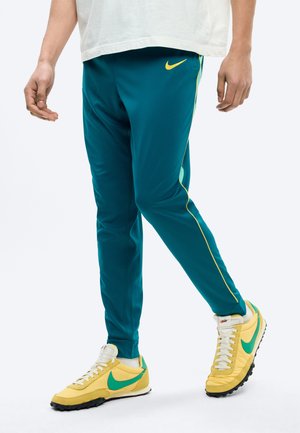 Persona con pantalones deportivos Nike color verde azulado con ribetes amarillos y zapatillas Nike amarillas con el logo verde, de pie sobre un fondo blanco.