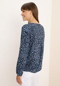 Blusa a maniche lunghe in un tessuto blu scuro con stampa astratta blu, scollatura rotonda e dettagli arricciati sulle spalle.
