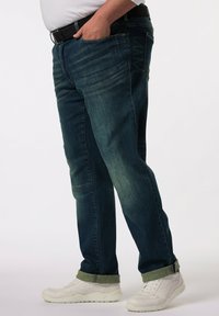 Tmavě modré džíny s efektem vyblednutí, rovného střihu, s ohrnutými nohavicemi, v kombinaci s bílými teniskami. U pasu je pásek.