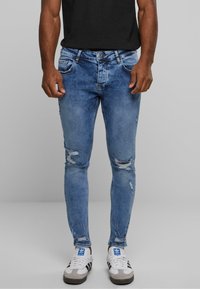Jeans ajustados de mezclilla azul con detalles desgastados. Cuenta con cinco bolsillos, cierre de cremallera y un aspecto desvaído. Combinados con zapatillas blancas.