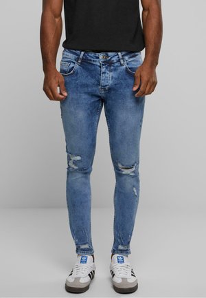 Jean skinny en denim bleu avec des détails déchirés. Comprend cinq poches, une fermeture éclair et un aspect délavé. Associé à des baskets blanches.