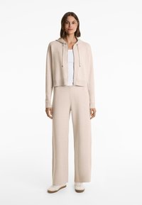 Donna in piedi che indossa un hoodie beige con zip, pantaloni a gamba larga, canotta bianca e sneakers bianche su uno sfondo bianco.
