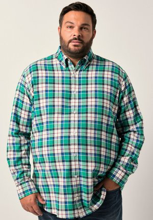 Man die staat met één hand in de zak, gekleed in een groen, wit en blauw geruit overhemd en donkere broek, tegen een effen achtergrond.