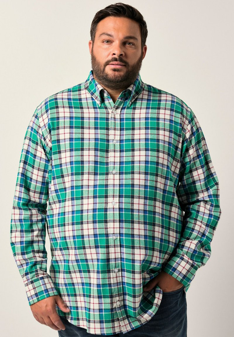 Uomo in piedi con una mano in tasca, che indossa una camicia a quadri verde, bianca e blu con bottoni e pantaloni scuri, su uno sfondo semplice.
