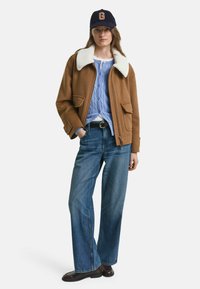 Hnedá bomberová bunda z vlnenej zmesi s bielym umelým kožúškom, skombinovaná so svetrom z modrého pleteného vzoru a širokými modrými džínsami. Čierne topánky.