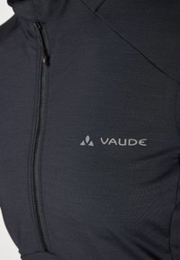 Veste noire en tissu texturé avec fermeture éclair sur le devant et le logo "Vaude" imprimé en gris sur le côté gauche.