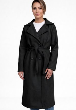 Kvinde iført en lang sort kåbe med bælte over en hvid top og blå jeans, der står mod en hvid baggrund.