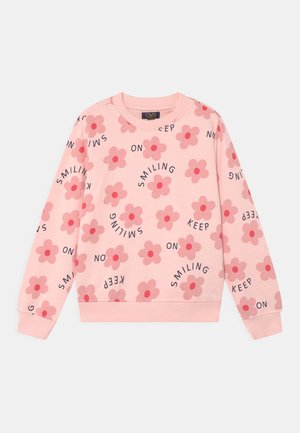 Lyserød sweatshirt med lange ærmer med blomsterprint og sort tekst "KEEP ON SMILING" spredt over stoffet.