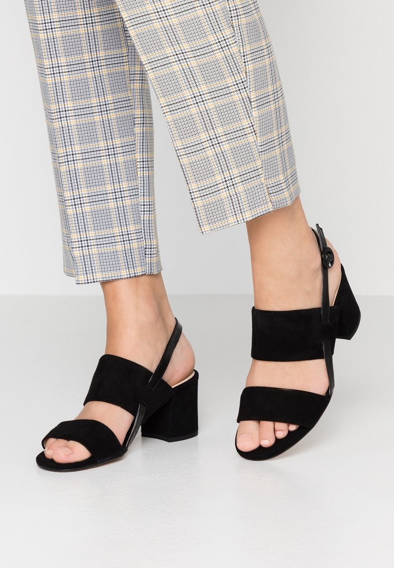 Sandales en daim noir avec bout ouvert, deux larges brides sur le pied et un talon bloc. Comprend une bride arrière avec un détail à bouton.